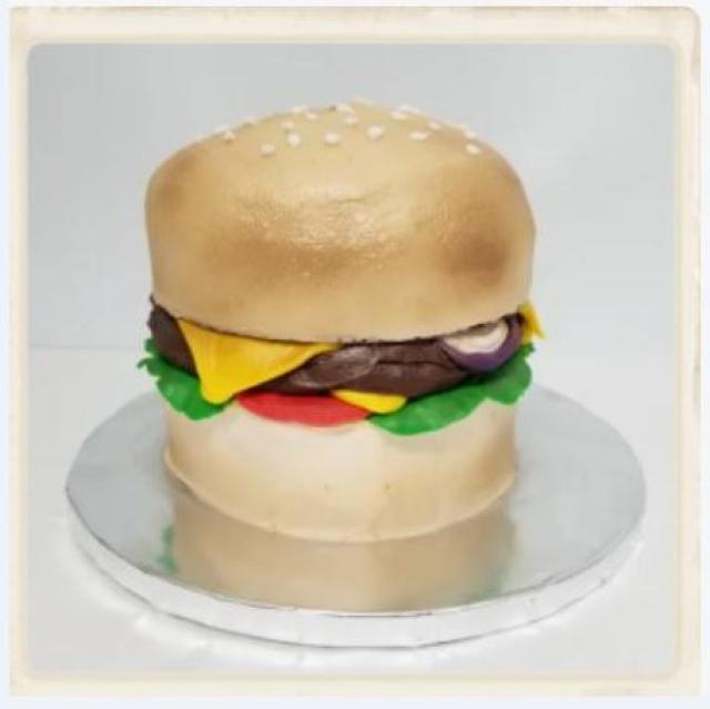 A cake made to look like a hamburger, complete with "cheese," "lettuce," "tomatoes," "onions," and a "sesame seed bun."," 